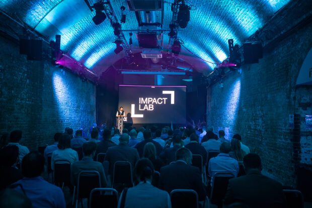 Audience watching speaker on stage with Impact Logo on screen behind him