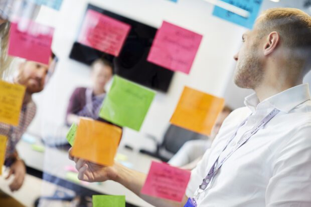 Man placing a sticky note on glass wall of a meeting room with colleagues in the background